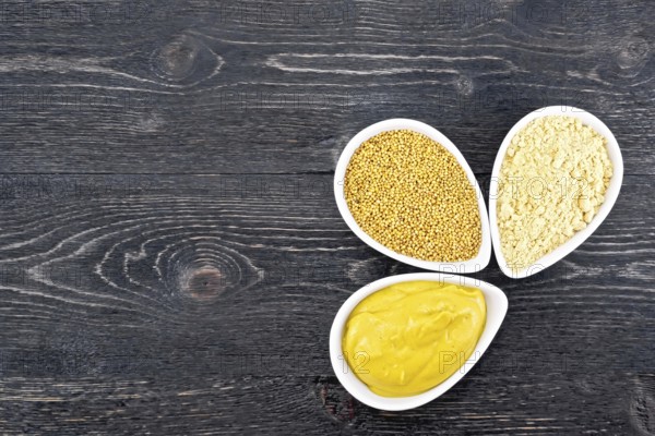Mustard sauce, seeds and mustard powder in three saucepans on a wooden plank background