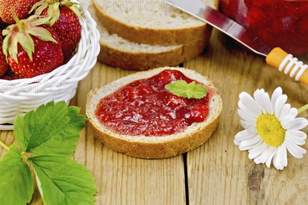 Bread with strawberry jam, a jar of jam, knife, strawberries in a wicker basket, chamomile on a wooden boards background