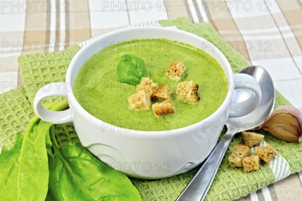 Green soup puree in a white bowl with croutons and spinach leaves, spoon on a napkin on the background fabric