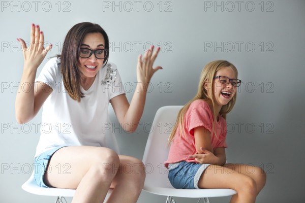 Happy mother and her little daughter sitting on the chairs by the grey wall. Portrait of mother and daughter with eyeglasses