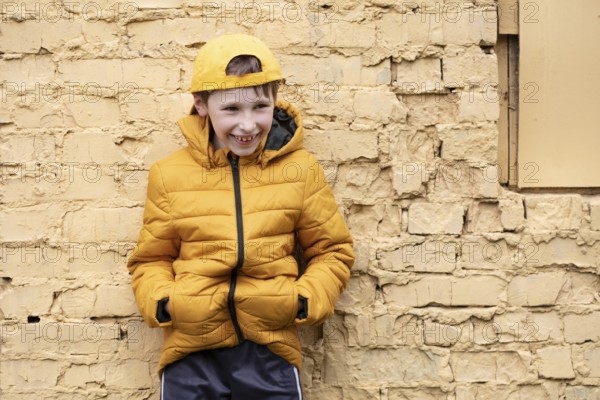 A child on the street A cheerful boy in yellow clothes and a baseball cap stands against a brick wall