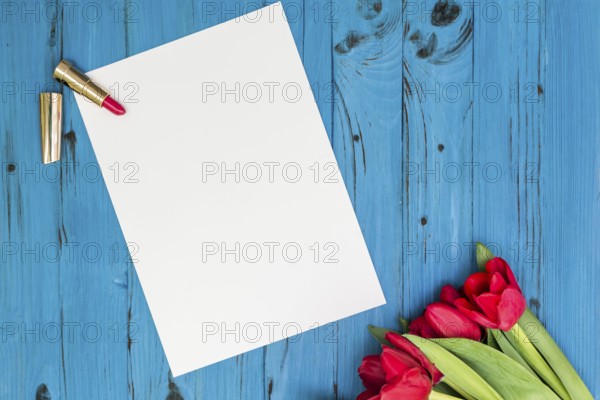 Red tulips, lipstick and a white sheet of paper for your greetings on the background of blue wooden board