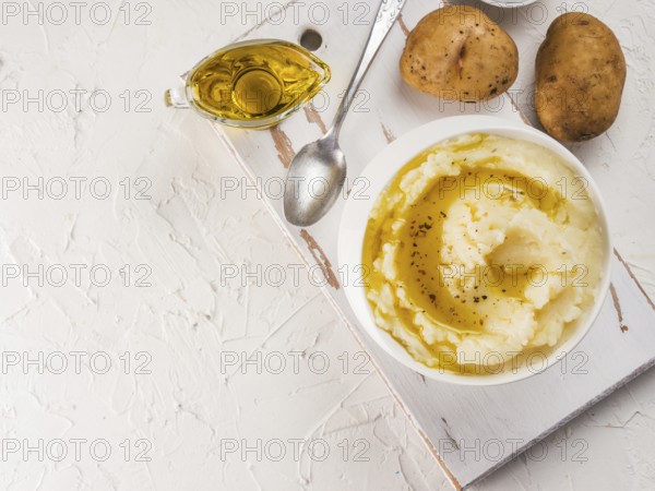 Mashed potatoes in white bowl on white cutting board, pepper and olive oil on white concrete background. Top view or flat lay. Copy space
