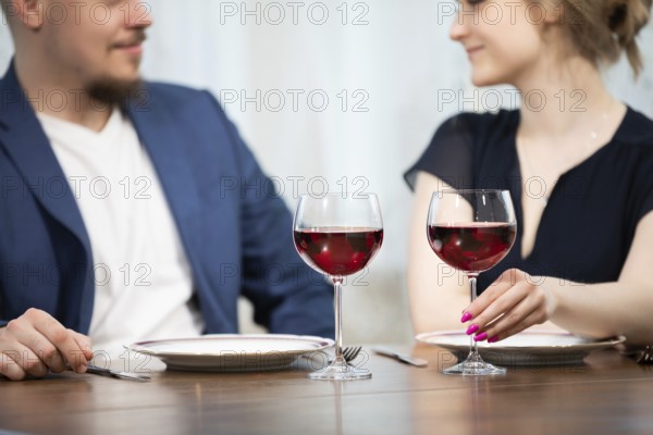A man and a woman sit at a table. Both are elegantly dressed. On the table are two plates and two glasses of red wine. The woman is holding one of the glasses