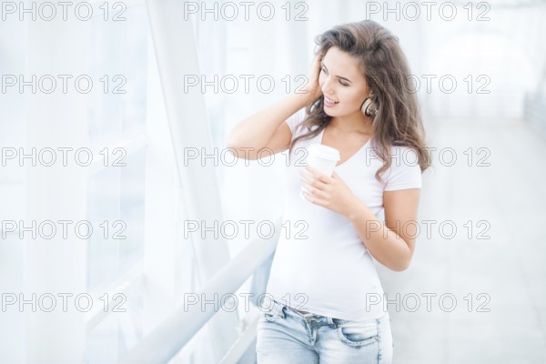 Happy young woman with music headphones standing on the bridge with a takeaway coffee mug, looking to the side against the urban backdrop