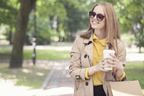 Young fashionable woman taking a coffee break after shopping and walking with a coffee to go in her hand against the backdrop of a green city park