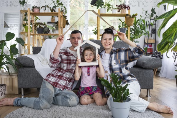 Traditional family with child are enjoying new home, sitting on the sofa and holding the roof. Mortgage, insurance and protection, buying and moving into your own home, green house with potted plant