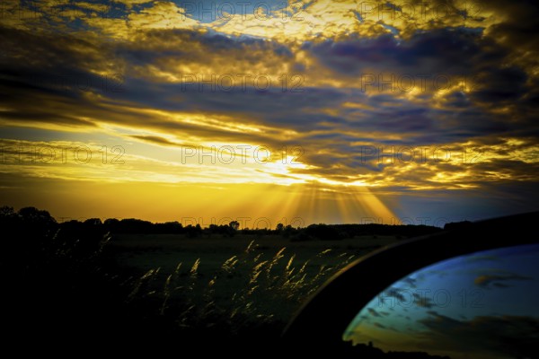 A dramatic sunset with yellow and blue tones, rays of light through clouds and a dark silhouette in the foreground and reflections in a car rear-view mirror, Polder 27798 Hude, Germany, Europe