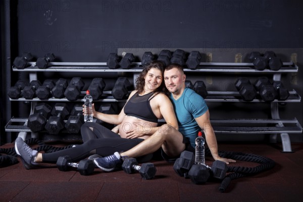 Pregnant woman goes for sports with her husband in gym for cross training. Couple athletes expecting baby while resting after training and exercises to strengthen health and muscles expectant mother