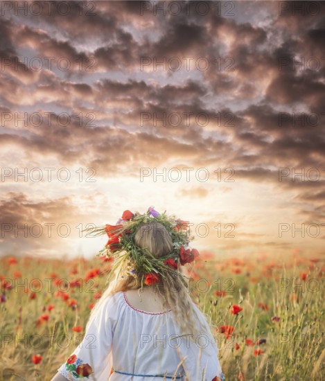 Pretty blonde girl in a dress with a pattern poppy and with a wreath on his head enjoying in a field with poppies. View from the back, copy space for inspired text
