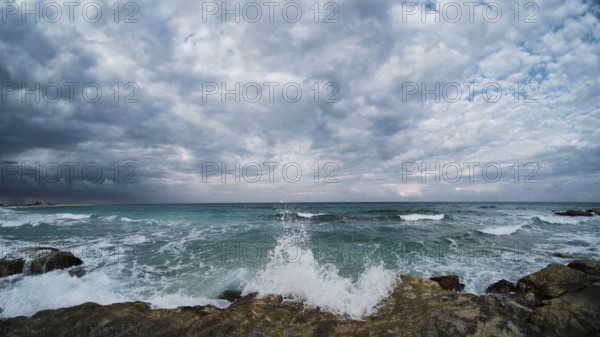 Waves of rushing sea are crashing over rocky coastline. Sea waves break upon rocks. Ultra-wide angle landscape with clouds and rushing sea