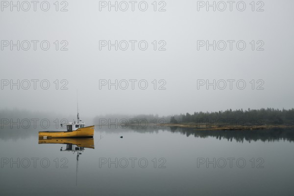 A commercial fishing boat is reflected in the still calm water of a protected bay as light fog begins to cover the shoreline in Lunenburg, Nova Scotia, Canada, North America