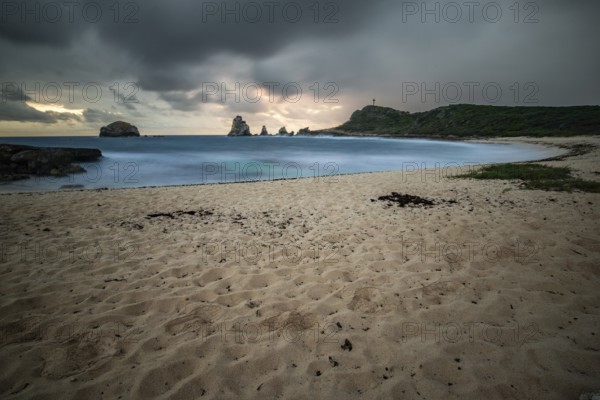 A secluded bay with a sandy beach in the morning. At the end of the beach a pointed rock formation extends into the sea, a stronger swell at a rainy sunrise creates a dramatic mood in a long exposure. Pointe des Chateau with view of Pointes des colibris on Grande Terre, Guadeloupe, French Antilles, Caribbean, North America