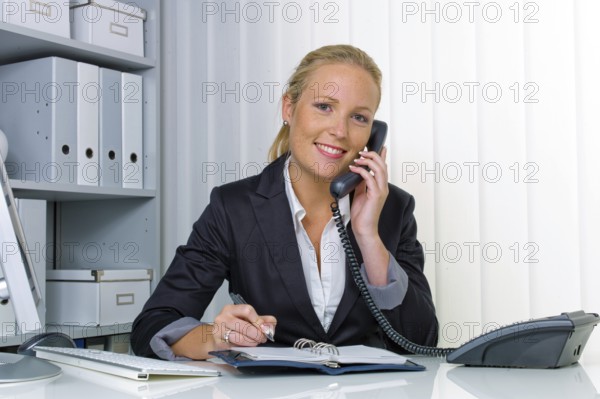 A friendly woman talks on the phone at her desk in the office and notes appointments in her calendar Austria