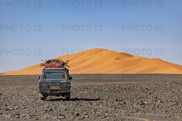 Off-road vehicle leads through a rocky plain in front of large sand dunes under a blue sky, By off-road vehicle through the Sahara desert in Algeria
