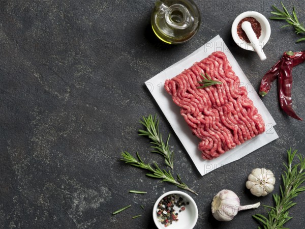 Fresh raw minced beef on backing paper and cutting board and ingredients over black cement background with copy space. Top view or flat-lay