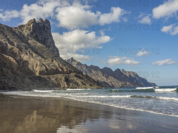 A sandy beach with waves, steep rocks and a clear blue sky with scattered clouds, tenerife, canary islands, spain