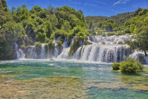 Waterfalls flow into an emerald green river, surrounded by dense forest and a bright blue sky, Krka National Park Lozovac Dalmatia Croatia