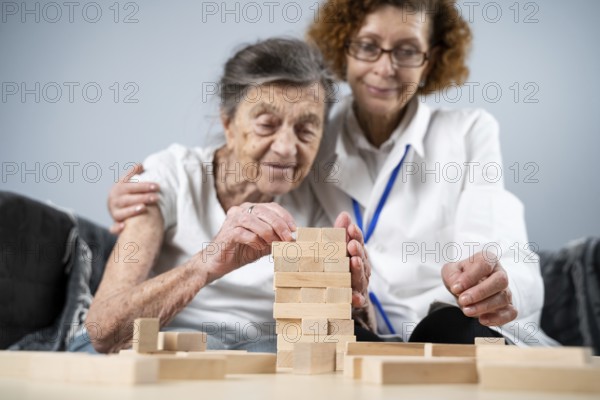 Senior woman practice skills, build wooden blocks, build tower and try not to let it fall, Jenga game. Old patient pull out block, place on top, support doctor during therapy dementia in house