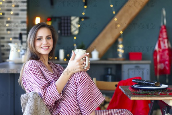 Happy young woman in plaid pajama holding jar smiling at camera in kitchen decorated for Christmas