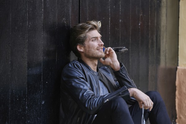 Young man with smartphone earphones sitting in the street black door