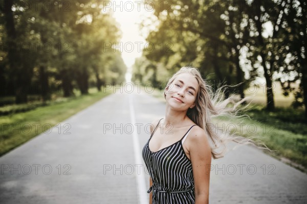 Woman standing on a road surrounded by trees, sunlight filtering through, creating a relaxed summer atmosphere