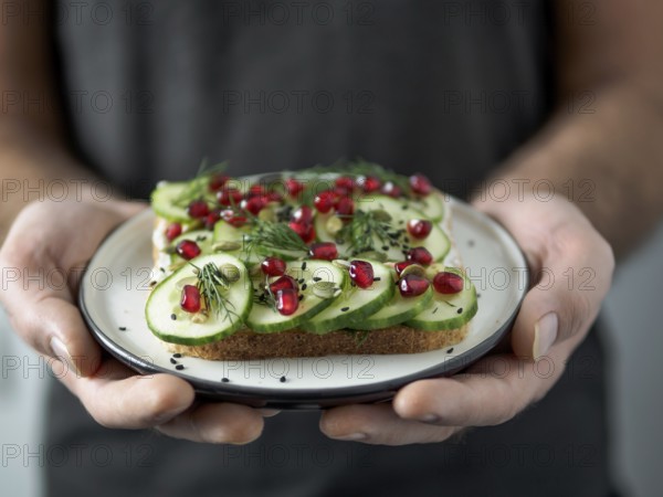 Hands takes plate with vegan sandwich. Healthy appetiezer - whole wheat bread toast with cucumber, pomegranate and black sesame. Vertical