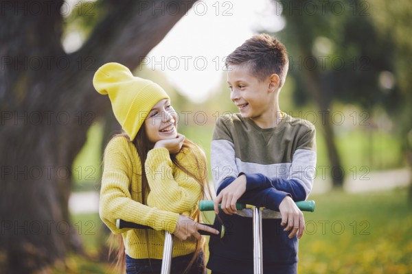 Twins 10 years old in bright clothes spend time actively together in autumn park riding kick scooters. Happy children. Eco transport. Kids posing in fall park. Brother and sister pre teens portrait