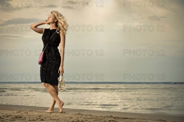 Fashionable young woman in dress with handbag paddling on sandy beach
