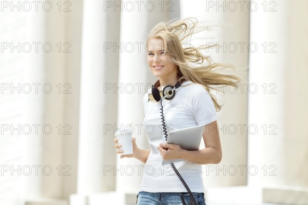 Beautiful young woman with vintage music headphones around her neck, holding a takeaway coffee and standing with wind in her hair against urban city background