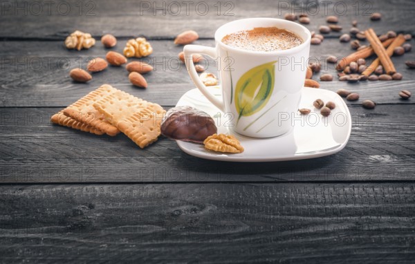 Cup of hot coffee on a plate, surrounded by tasty and healthy snacks, biscuits, almonds, walnuts and coffee beans, on an aged wooden table, in the morning light