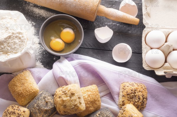 High angle view with a bag of flour, eggs, a rolling pin and freshly baked bread rolls, decorated with different healthy seeds, on a wooden black table