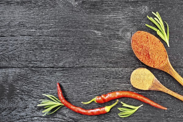 Frame of sprigs of fresh rosemary, pods of red hot pepper and two spoons with ground pepper on a wooden board background from above