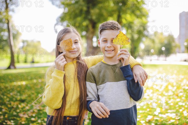 Happy Caucasian siblings enjoying in autumn day in park, sunny weather. Kids having fun holding fallen yellow leaves in nature. Funny twins in autumn park. Autumnal mood. Teenagers posing embracing