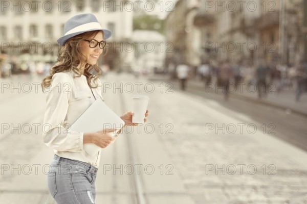Beautiful young woman with a disposable coffee cup drinking coffee, holding a tablet in her hands and crossing the street against an urban background