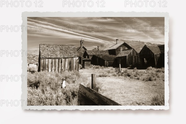 A dusty path between thorny undergrowth leads to wooden houses in the former gold mining town of Bodie. Photo in sepia tones with white jagged edges in the character of old photos, lying shadow. A dusty path between thorny shrubs leads to wretched wooden houses of the former gold mining town Bodie. Photo in sepia tone with white jagged edges