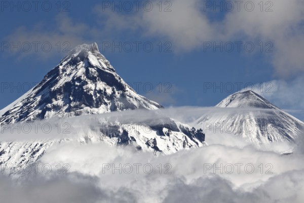 Stunning landscape view of snowy top of rocky volcanoes cones of Kamen Volcano and active Klyuchevskoy Volcano above clouds. Russian Far East, Kamchatka Peninsula, Klyuchevskaya Group of Volcanoes