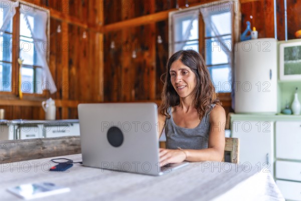 Smiling freelancer woman working on vacation inside a wooden mountain chalet, using a laptop sitting at the table