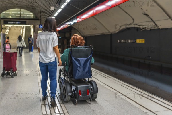 Rear view of a disabled caucasian adult woman in wheelchair and asian friend walking along metro station
