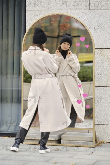 A woman stands outdoors, looking at her reflection in a modern mirror while wearing a coat and a beanie