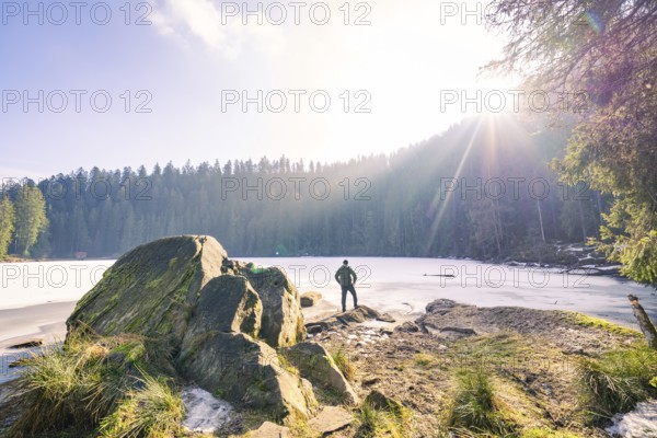 Man standing on rocks in front of a frozen lake, surrounded by forest, sunlight illuminates the landscape, Glaswaldsee, Bad Rippoldsau-Schapbach, district of Wolfach, Black Forest, Germany, Europe