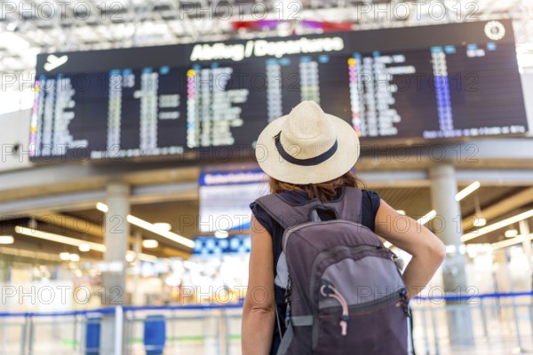 A woman wearing a straw hat and a backpack is standing in front of a large airport display. She is waiting for her flight, possibly at an airport