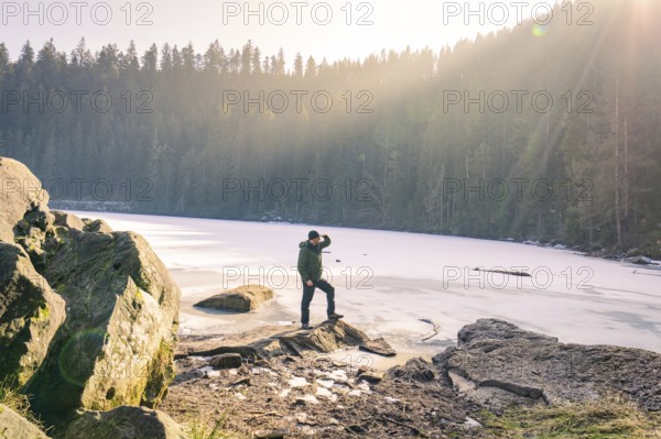 Person standing on the shore of a frozen lake surrounded by forest and sunbeams, Glaswaldsee, Bad Rippoldsau-Schapbach, district of Wolfach, Black Forest, Germany, Europe