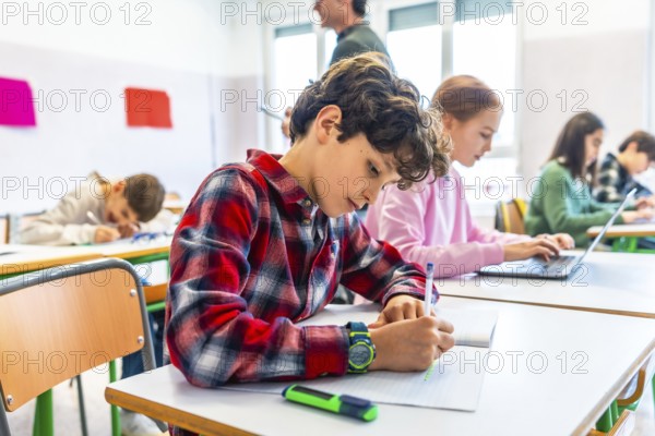 Elementary school students are diligently working at their desks, some writing in notebooks and others using laptops, showcasing a modern learning environment
