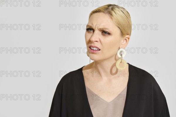A woman in a black outfit stands against a light gray background, displaying a confused expression. Her large statement earrings and hairstyle emphasize her thoughtful demeanor, creating contrast
