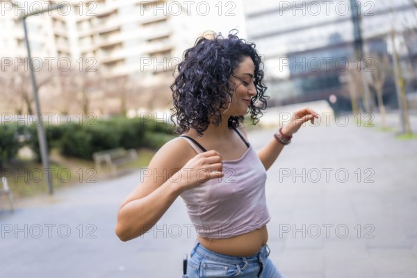 Curly haired hispanic woman joyfully dancing in an urban setting, embracing the sunny day with vibrant energy and carefree movement
