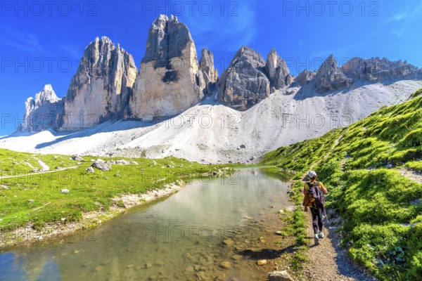 Tourist hiking along a scenic trail by a crystal clear alpine lake, with the stunning tre cime di lavaredo peaks in view