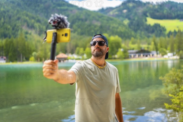 Content creator filming a video with a smartphone and external microphone at lake dobbiaco in the italian dolomites during summer