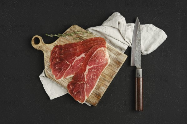 Overhead view of jamon iberico cured pork meat on a wooden cutting board, accompanied by a sharp knife. A soft cloth adds texture, while a sprig of thyme enhances the presentation, suggesting a culinary preparation.