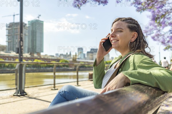 A cheerful woman sitting on a bench by the riverside, talking on her phone, with modern city buildings and blooming trees in the background on a sunny day